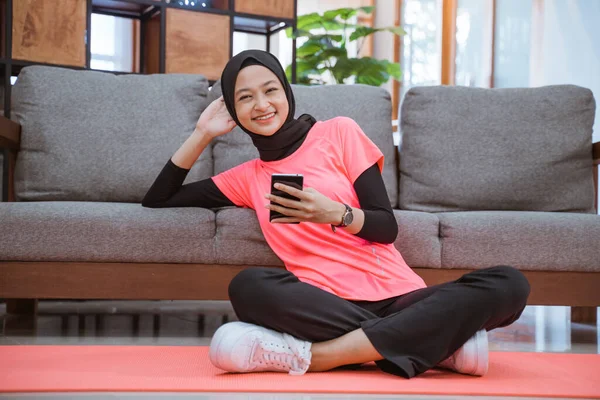 Una chica en una ropa de gimnasio hijab sonriente sosteniendo un teléfono celular mientras está sentada relajada en el suelo —  Fotos de Stock