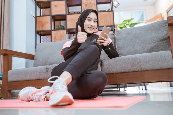 Žena na sobě hidžáb sportovní oblečení s úsměvem na kameru při použití mobilu s palci nahoru, zatímco sedí uvolněně — Stock fotografie
