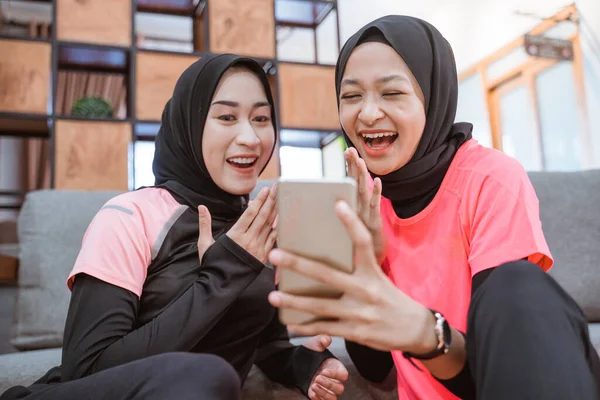 Dos mujeres que usan ropa deportiva hijab ríen mientras leen mensajes juntos en un teléfono celular — Foto de Stock