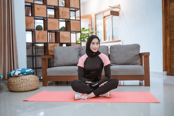 Una mujer con una ropa deportiva de hijab se sienta en el suelo sobre un colchón sobre el fondo del sofá —  Fotos de Stock