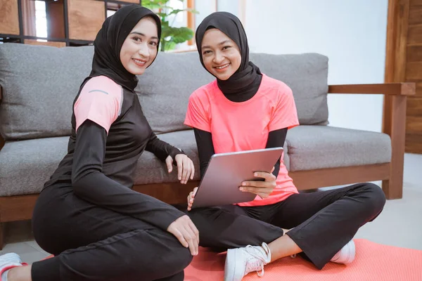 Dos mujeres jóvenes que usan ropa deportiva de hijab sonriendo mientras se relajan en el suelo mientras usan una tableta digital —  Fotos de Stock