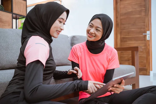 Duas mulheres vestindo roupas esportivas hijab enquanto estão sentadas conversando casualmente no chão enquanto usam um tablet digital — Fotografia de Stock