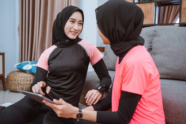 Dos mujeres usando hijab sportswear sentarse a charlar en el suelo mientras se utiliza una tableta digital juntos —  Fotos de Stock