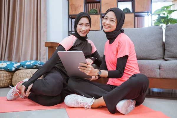 Dos mujeres musulmanas que usan hijab sportswear sonríen mientras se relajan en el suelo mientras usan una tableta digital —  Fotos de Stock
