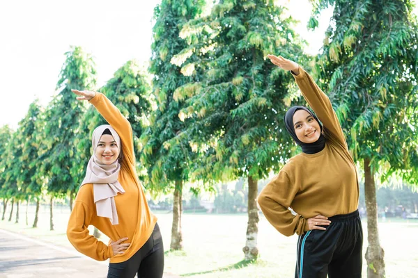 Dua gadis Muslim dalam kerudung meregangkan otot-otot mereka dengan mengangkat tangan mereka untuk berolahraga di luar ruangan — Stok Foto