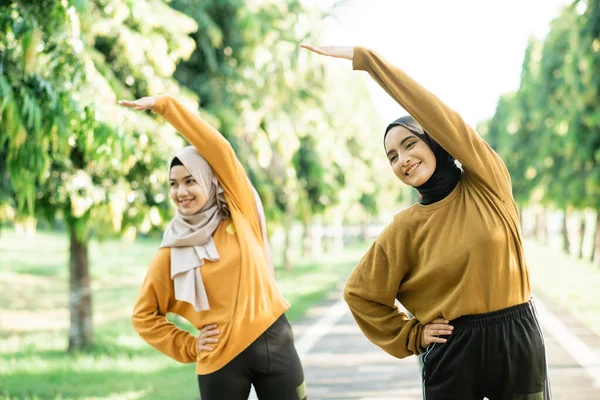 Dos chicas musulmanas asiáticas en pañuelos para la cabeza estiran sus músculos levantando las manos para hacer ejercicio fuera de la habitación —  Fotos de Stock
