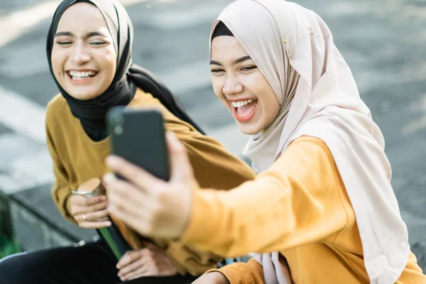 Dos chicas que usan hijab riendo cuando buscan vídeo junto con el teléfono inteligente por la tarde — Foto de Stock