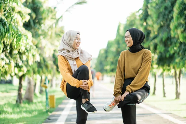 Dua gadis berkerudung meregangkan otot kaki mereka dengan mengangkat dan memegang kaki bengkok dengan tangan mereka sebelum jogging — Stok Foto