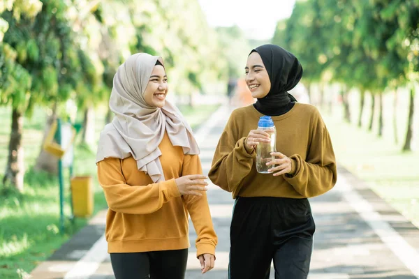 Дві азіатські мусульманські дівчата насолоджуються спортом разом, спілкуючись і тримаючи питну воду з пляшкою — стокове фото