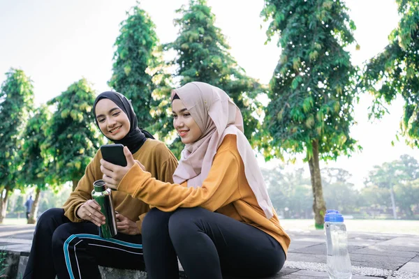 スマートフォンを使ったイスラム教徒の10代の少女2人とスポーツをした後の飲料水ボトルを持って — ストック写真