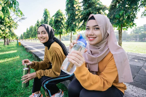 Leende två muslimska tonårsflickor dricka vatten med hjälp av flaskor efter sport tillsammans på eftermiddagen — Stockfoto