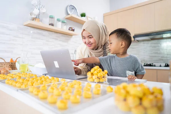 Ibu muslim melihat laptop sambil membuat kue dengan anaknya di dapur — Stok Foto