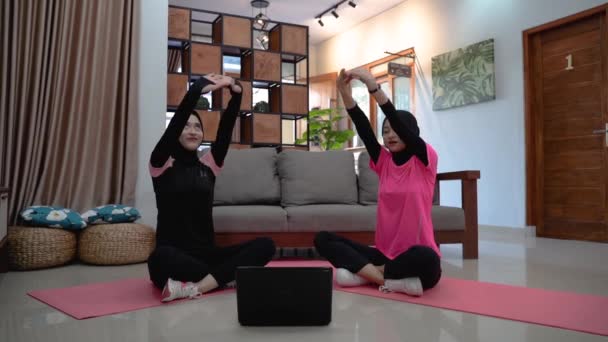 Две женщины в хиджабе сидят на полу со скрещенными ногами, прислонившись к бокам и подняв руки, пока разогреваются. — стоковое видео