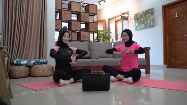 Dwie kobiety ubrane w hidżabską odzież sportową siedzą na podłodze ze skrzyżowanymi nogami, pochylone do boku i ręce w górze, rozgrzewając się. — Wideo stockowe