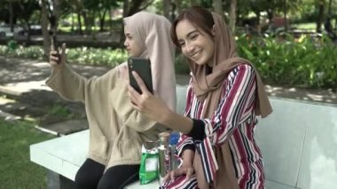 İki kız tesettür takıyor, gülümsüyor ve akıllı telefon kamerasıyla selfie çekerken akıllı telefon tutuyorlar.