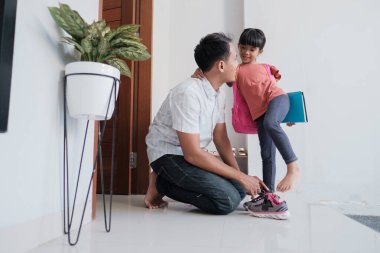 Baba okula gitmeden önce kızına ayakkabı giymesinde yardım ediyor.