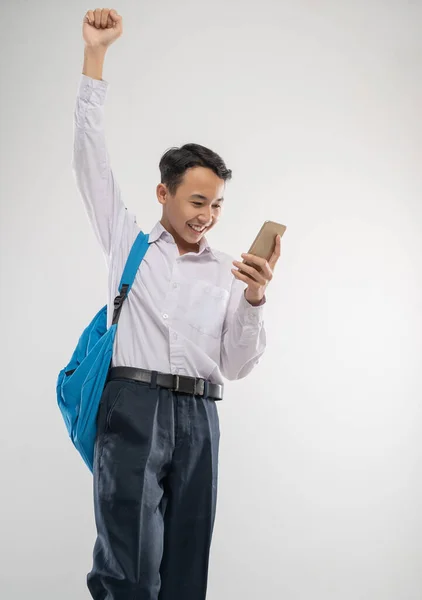 A excited boy wears a school uniform using a handphone while raises his hand and carries a backpack — Stock Photo, Image