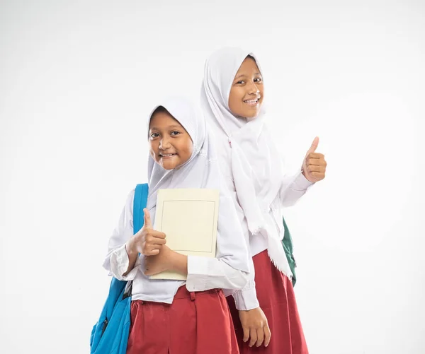 Sorridendo due ragazze velate che indossano uniformi scolastiche elementari con i pollici alzati che trasportano zaini e un libro — Foto Stock