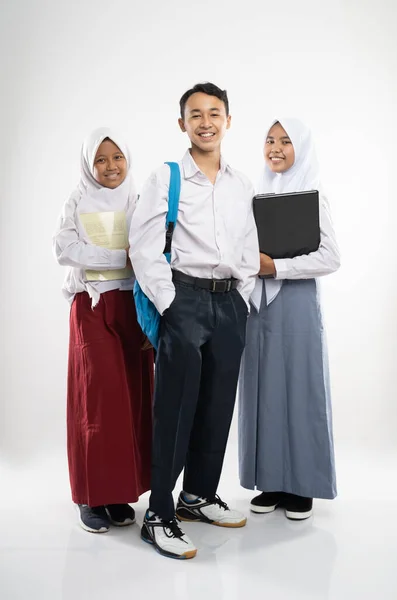 Three Indonesian teenagers wearing school uniforms smile at the camera with a backpack, a book, and a laptop computer — Stock Photo, Image