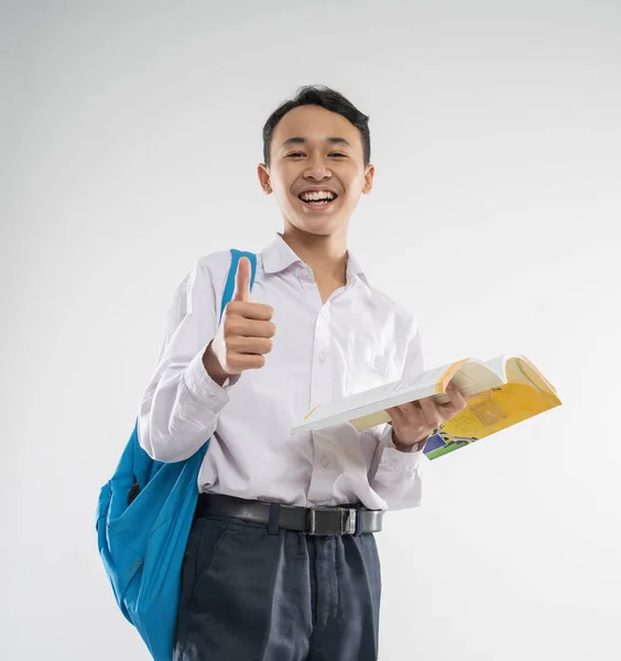 Un niño vistiendo un uniforme de secundaria sonríe mientras sostiene un libro con los pulgares hacia arriba —  Fotos de Stock