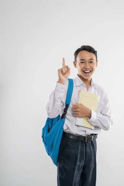 Um menino de uniforme do ensino médio sorrindo com o dedo apontando quando carregando um livro e uma mochila com copyspace — Fotografia de Stock