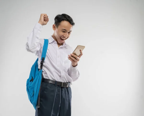 A boy wearing a junior high school uniform looking at a cellphone with an excited gesture with a backpack — Stock Photo, Image
