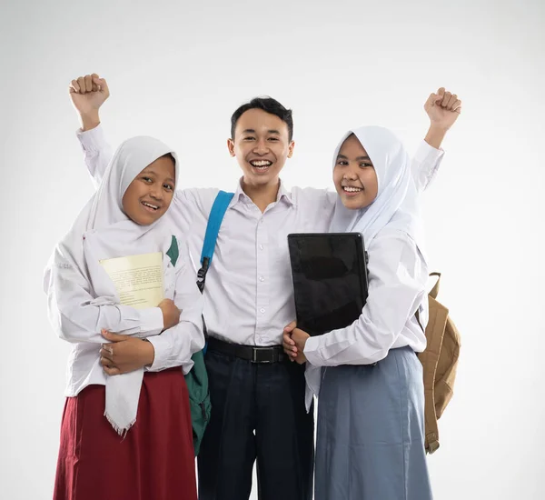 Três adolescentes usando uniformes escolares sorriem para a câmera com uma mochila, um livro e um computador portátil — Fotografia de Stock