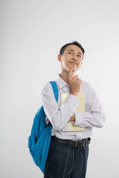 Een jongen in junior middelbare school uniform glimlachend omhoog kijkend bij het denken gebaar bij het vasthouden van een boek en een rugzak met copyspace — Stockfoto