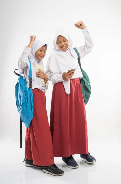 Dua gadis bercadar mengenakan seragam sekolah dasar menggunakan ponsel bersama-sama dengan ransel — Stok Foto