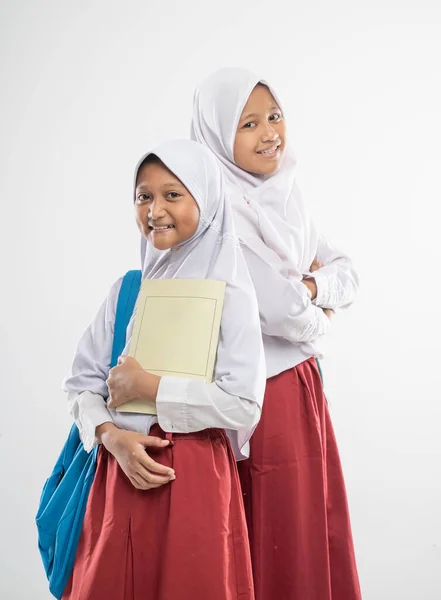 Dua gadis berkerudung Asia yang mengenakan seragam sekolah dasar berdiri satu sama lain sambil membawa tas ransel dan buku — Stok Foto