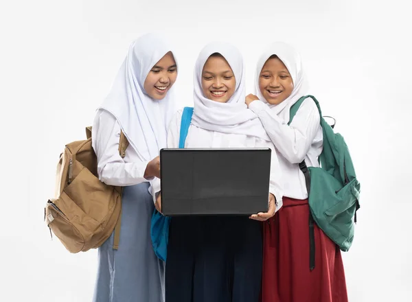 Három ázsiai lány fátyolban iskolai egyenruhában mosolyogva áll egy laptop segítségével, miközben hátizsákokat cipelnek. — Stock Fotó