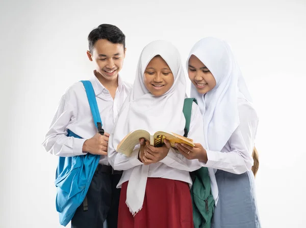 Két fátyolos lány és egy fiú középiskolai egyenruha mosolyog, amikor együtt olvasnak egy könyvet, miközben hátizsákot cipelnek. — Stock Fotó