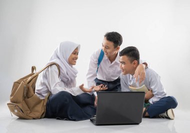 Ortaokul üniformalı üç genç birlikte dizüstü bilgisayar kullanırken yerde oturup sohbet ediyorlar.