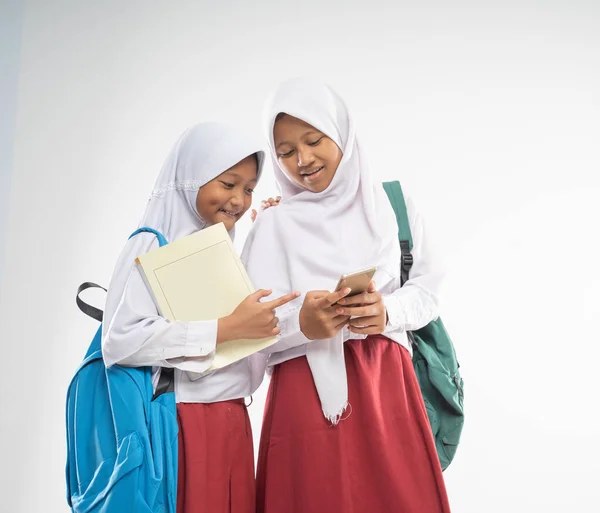 Dos chicas veladas con uniformes de escuela primaria mientras usan un teléfono móvil junto con una mochila y un libro —  Fotos de Stock