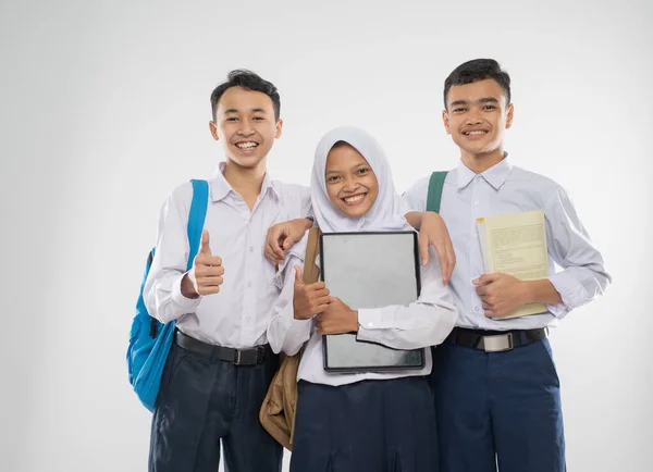 Tres adolescentes en uniformes de secundaria sosteniendo una computadora portátil con pulgares hacia arriba cuando llevan una bolsa de la escuela y un libro —  Fotos de Stock