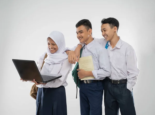 Tiga remaja dengan seragam SMP tersenyum menggunakan komputer laptop bersama sambil membawa tas ransel dan buku — Stok Foto