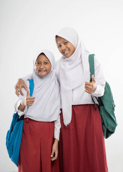 Dos chicas veladas en uniformes de escuela primaria con pulgares hacia arriba mientras llevan una mochila y un libro —  Fotos de Stock