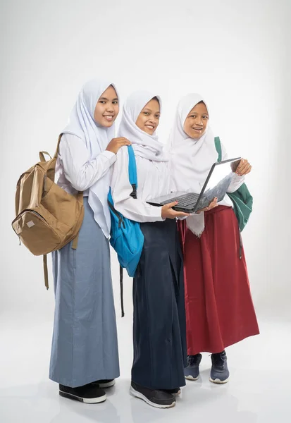 Tre leende flickor bär slöja i skoluniformer med hjälp av en bärbar dator tillsammans med skolväskor — Stockfoto