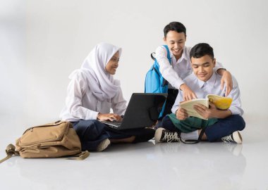Orta okul üniformalı üç gencin dizüstü bilgisayar ve kitap kullanarak yerde oturup birlikte ders çalışmasını izlemek.