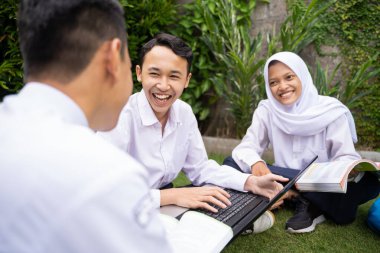 Okul üniformalı üç genç çimlerin üzerinde otururken dizüstü bilgisayar ve kitap kullanarak ders çalışıp sohbet ediyorlar.