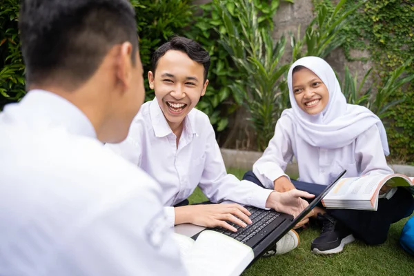 Tre teenagere i skoleuniformer studerer sammen og chatter med en bærbar computer og en bog, mens de sidder på græsset - Stock-foto