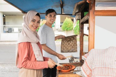 İki satıcı yemek tezgahlarında yemek hazırlıyor. Geleneksel tavuk satayı satıyor.