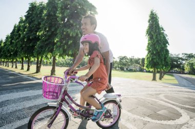 Baba kızına parkta bisiklet sürmeyi öğretiyor.