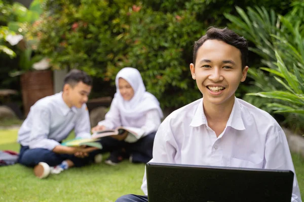 Seorang remaja berseragam sekolah tersenyum sambil menggunakan laptop yang sedang duduk di rumput — Stok Foto