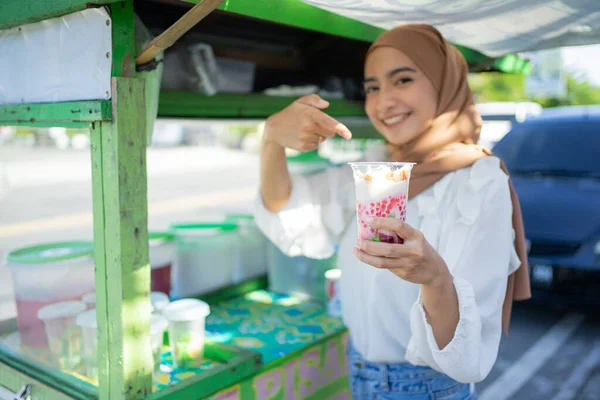 A pretty girl in veil of a es campur seller with her finger pointing at a plastic cup of delicious es campur — Stock Photo, Image