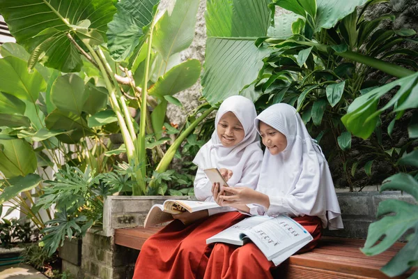 Dua anak mengenakan kerudung berseragam sekolah menggunakan ponsel dan buku sambil belajar bersama — Stok Foto