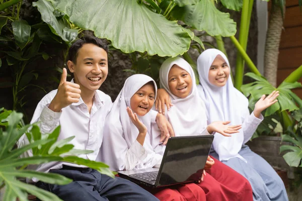 Four Asian teenagers in school uniforms smile at the camera while using a laptop — Stock Photo, Image