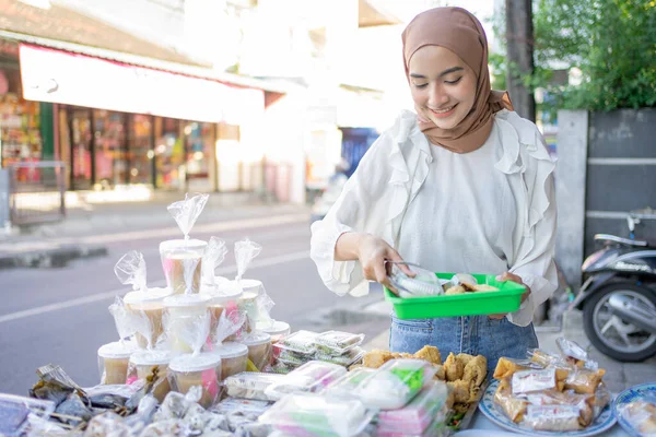 A beautiful girl in veil holding a plastic tray chooses various types of fritters to buy — Stock Photo, Image