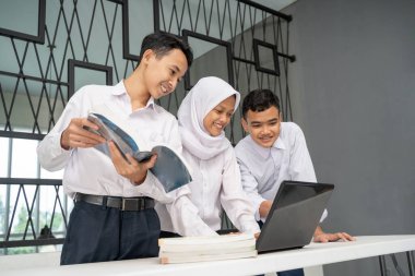 Üç Asyalı genç, bir dizüstü bilgisayar ve birkaç kitap kullanarak okul üniformalarıyla birlikte okuyorlar.