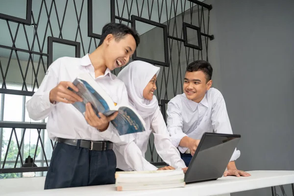 Three Asian teenagers study together in school uniforms to chat while using a laptop and several books — Stock Photo, Image